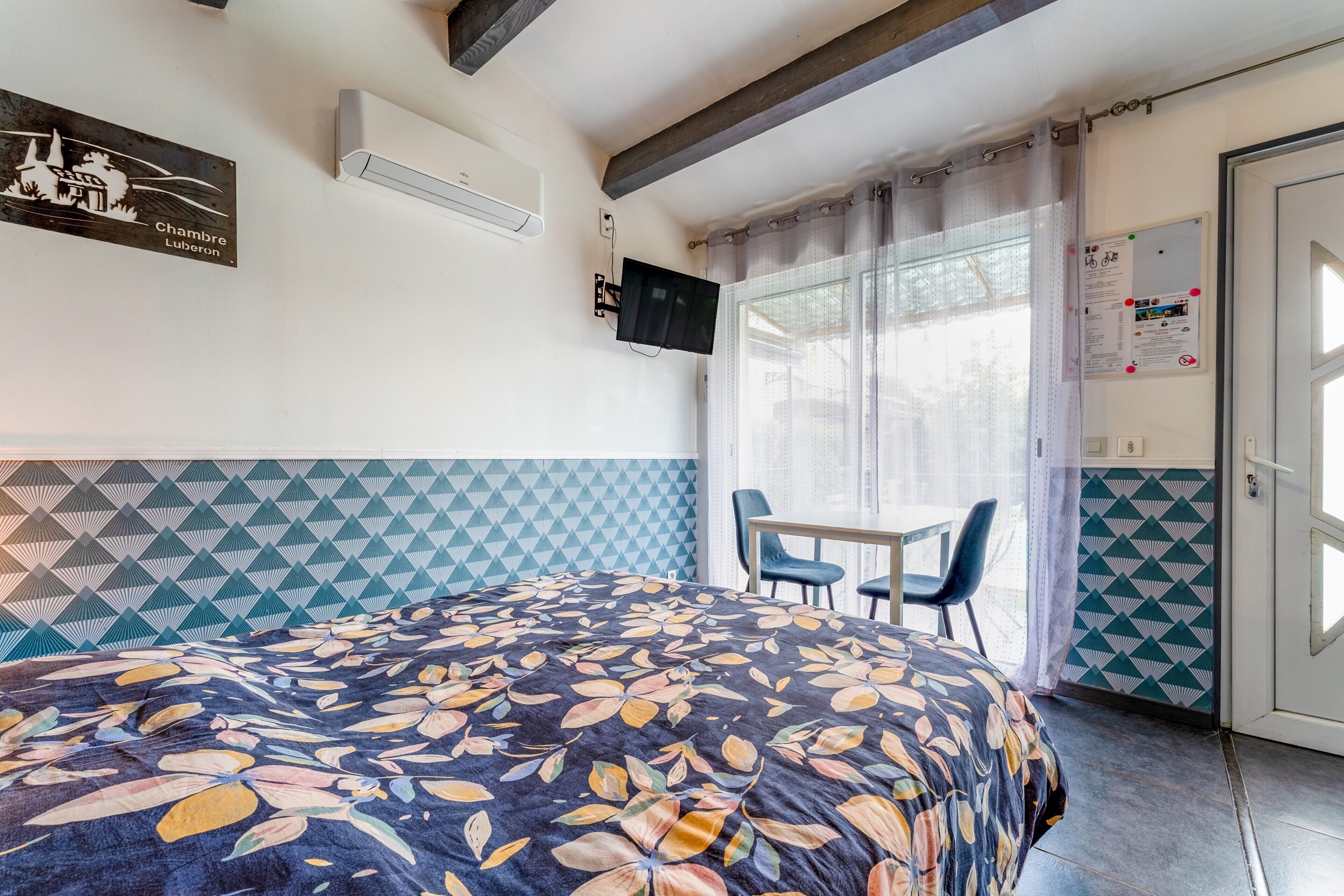 Una habitación de hotel con cama, televisión, mesa y sillas, una puerta y una ventana con cortinas.