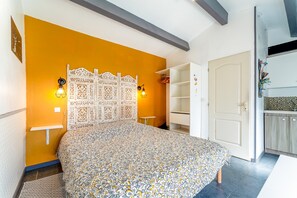 A bedroom with a patterned bedspread, a decorative headboard, two wall-mounted lights, a white wardrobe, and a door leading to a kitchen area.