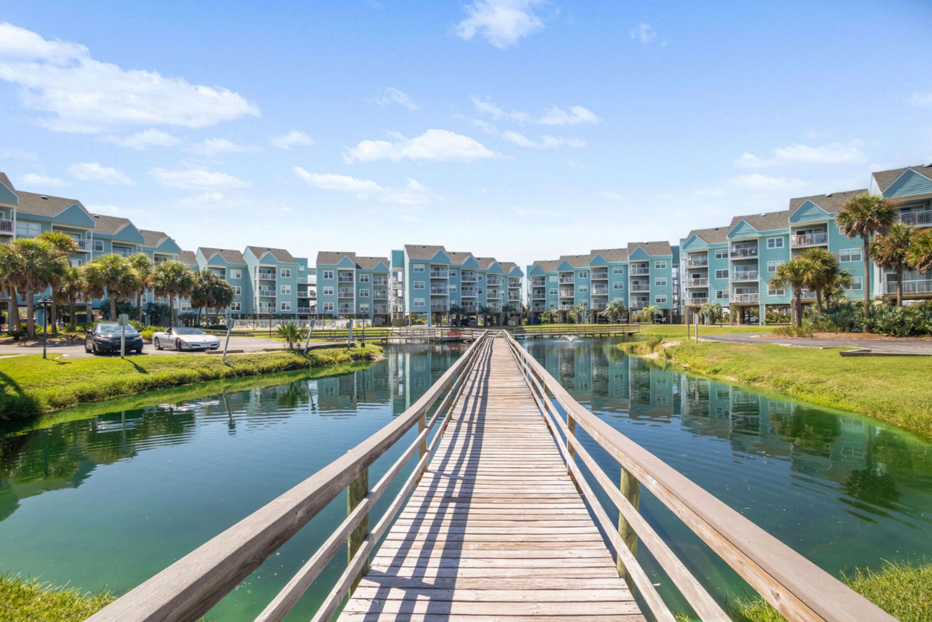 A wooden boardwalk leading to a row of blue buildings with balconies, over a calm water body, with cars parked nearby.
