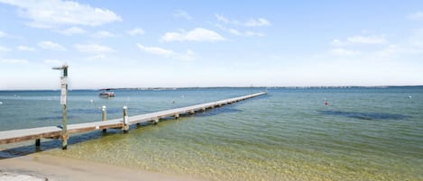 Una playa con un muelle de madera que se adentra en el agua, aguas claras y poco profundas, y una costa distante.