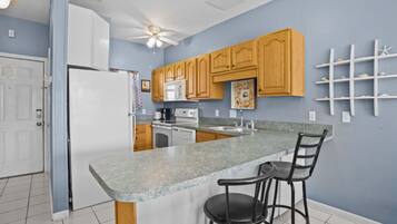 A kitchen with a granite countertop, wooden cabinets, a refrigerator, a microwave, a stove, and a ceiling fan.