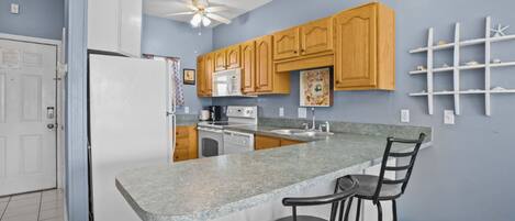 A kitchen with a granite countertop, wooden cabinets, a refrigerator, a microwave, a stove, and a ceiling fan.