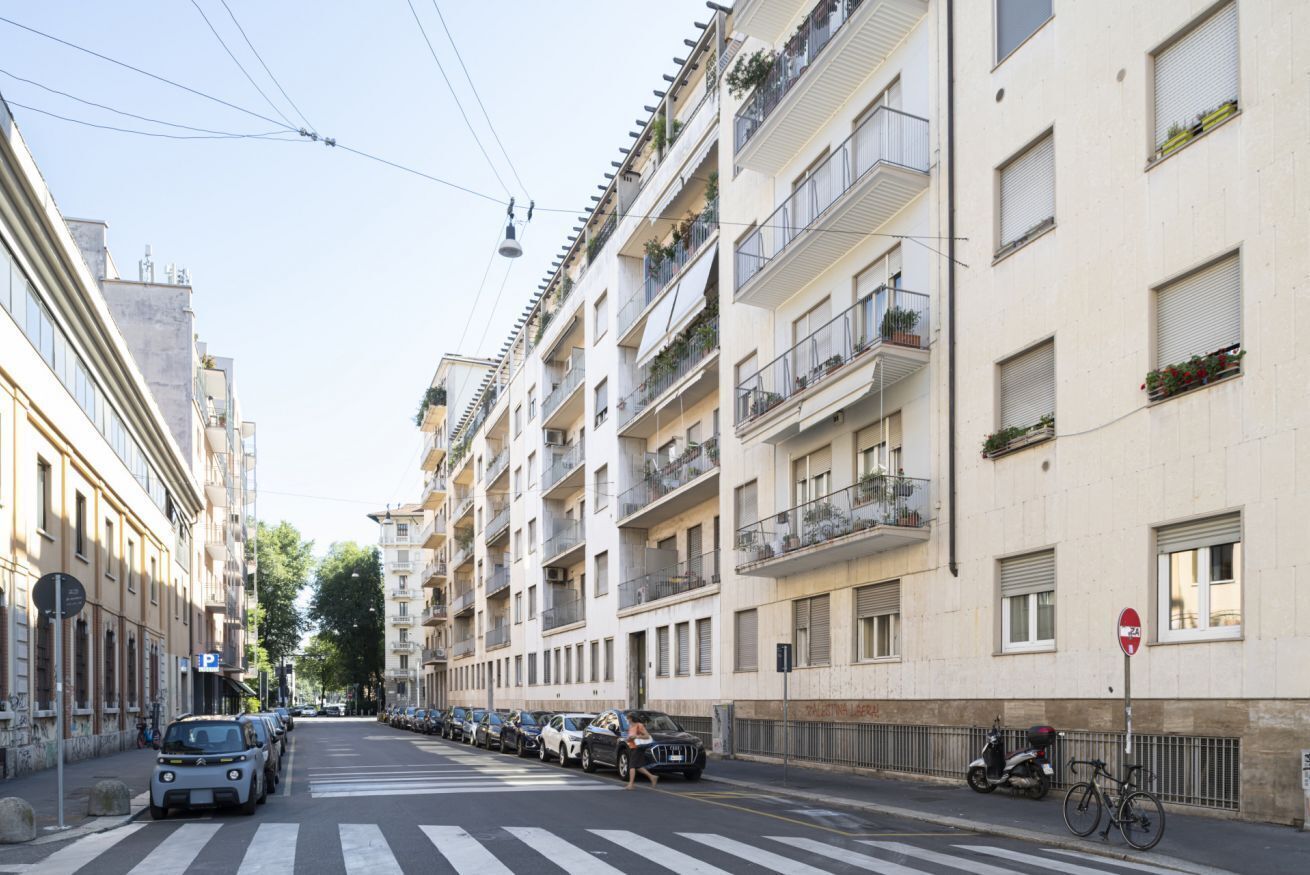 A street view with a row of multi-story residential buildings, a pedestrian crossing, parked cars, and a bicycle.