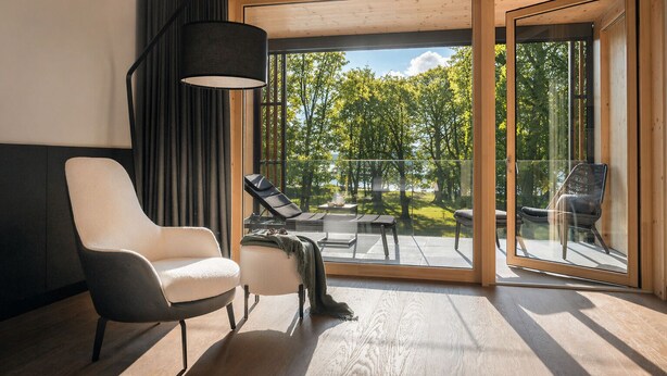 A modern room with a large sliding glass door leading to an outdoor area with a view of trees and a body of water. There are two chairs and a table inside the room.