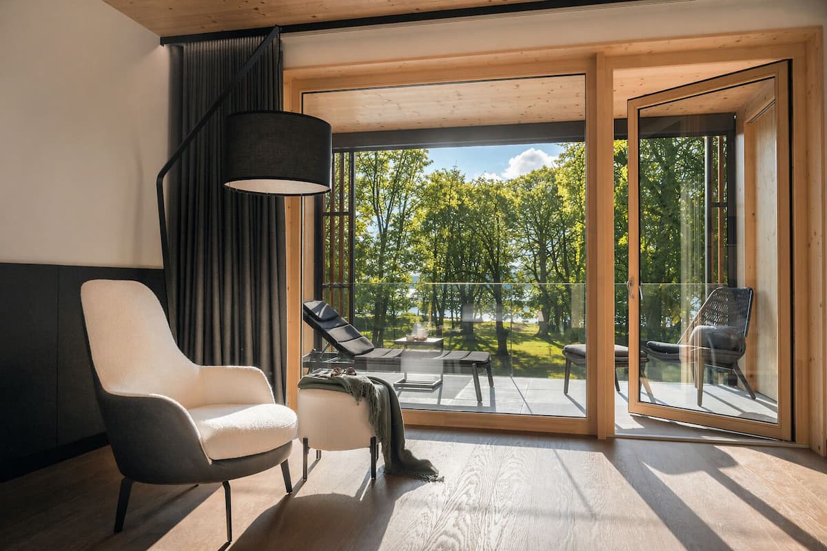 Ein modernes Zimmer mit einem großen Schiebeglasfenster, das zu einem Außenbereich mit Blick auf Bäume und Gewässer führt. Im Zimmer befinden sich zwei Stühle und ein Tisch.