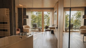 A modern bathroom with a large bathtub, a wooden floor, and a view of trees through large windows.