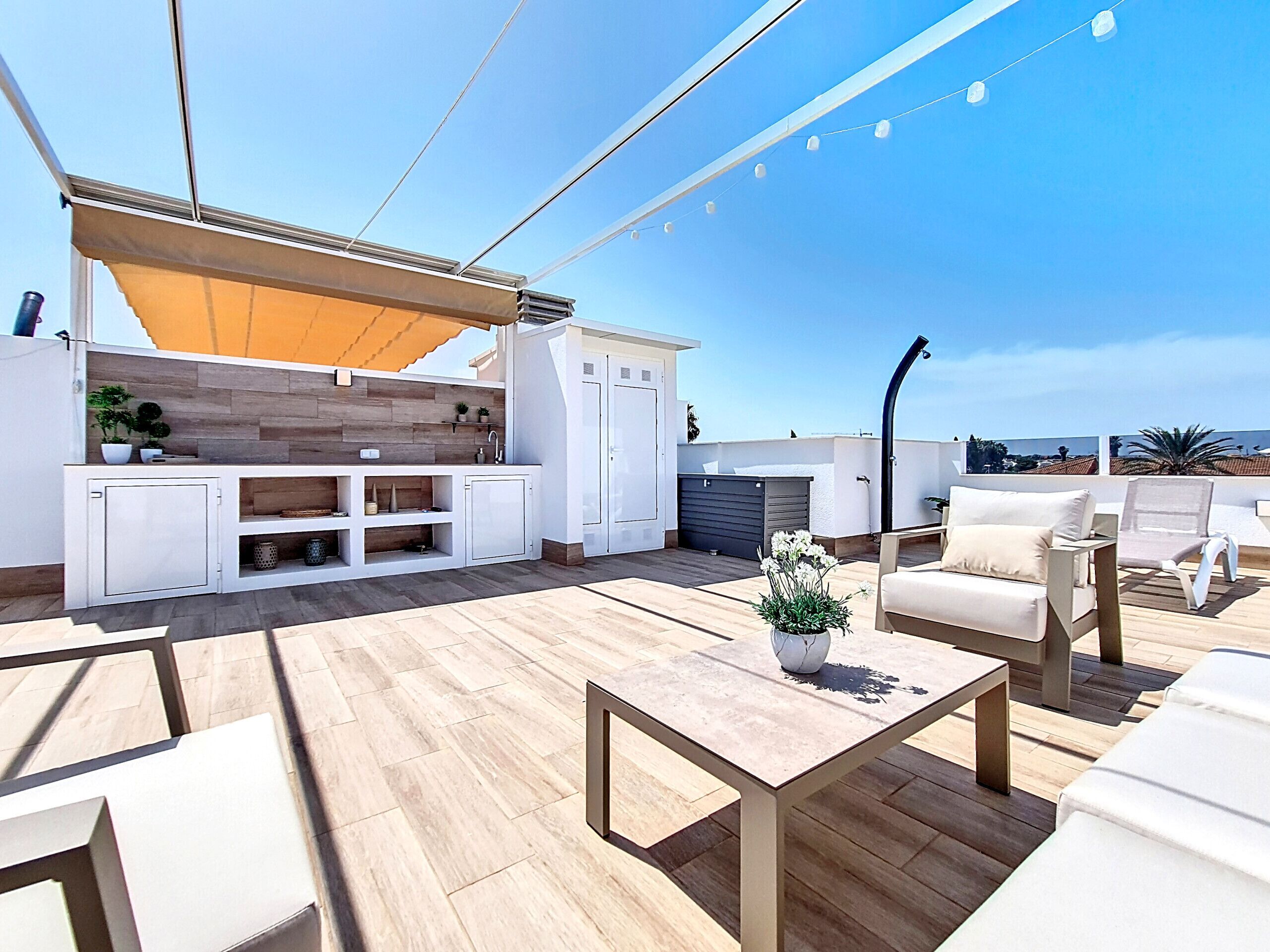 A rooftop terrace with a wooden deck, outdoor seating, a small kitchen area, and a view of the cityscape.