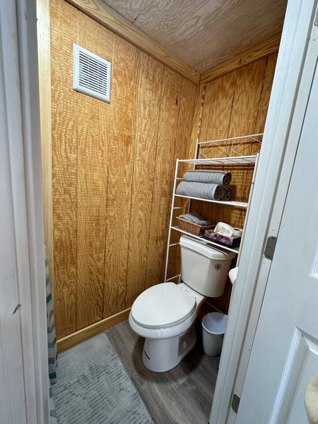 Et kompakt badeværelse med trævægge, et hvidt toilet, en metalkapel med håndklæder og en lille udluftning i væggen.