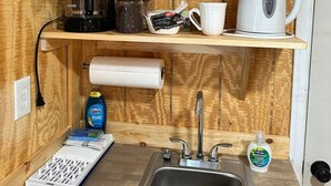 Cocina pequeña con un lavamanos, una cafetera, un hervidor y un rollo de papel higiénico sobre una repisa de madera.