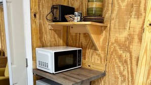 Una kitchenette compacta con un microondas blanco sobre un refrigerador pequeño, un estante de madera arriba y una silla amarilla en la esquina.