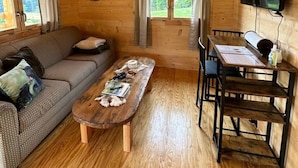 A cozy cabin interior with a wooden floor, a sofa, a coffee table, a dining table with chairs, and a TV mounted on the wall.