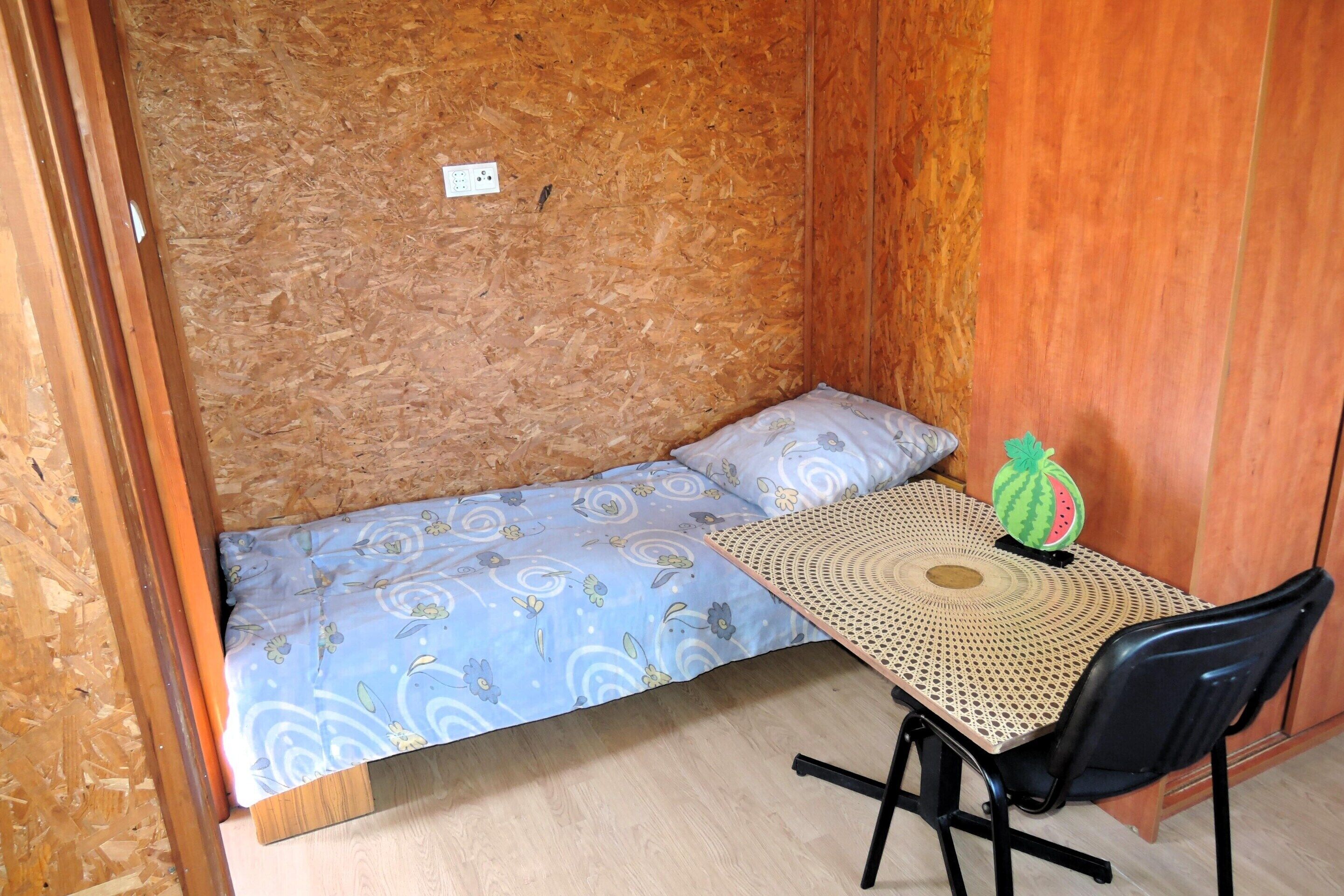 Ein kleines Zimmer mit einem Bett, einem Tisch mit einer gewebten Tischdecke und einem Stuhl.