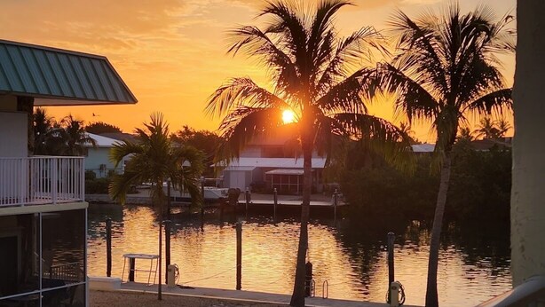 A waterfront property with a balcony overlooking a body of water, palm trees, and a building with a sloped roof. - Anchors Away- Beautiful condo w/ boat slip, boat ramp & Pool (Marathon)
