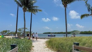 A sandy beach path with palm trees, a boat on the water, and people walking. - Anchors Away- Beautiful condo w/ boat slip, boat ramp & Pool (Marathon)