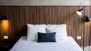 Una cama con sábanas blancas y una manta azul oscuro, flanqueada por paneles de madera y dos lámparas de pared.