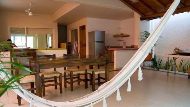 Un comedor con mesa y sillas de madera, una hamaca y una cocina al fondo.
