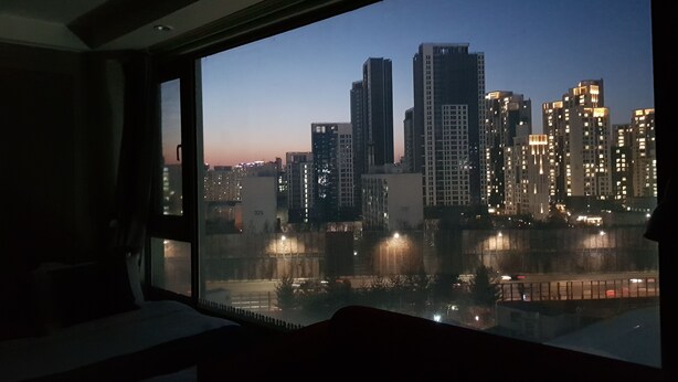 Vista da janela de um horizonte urbano ao entardecer, com edifícios iluminados e um corpo d'água em primeiro plano.