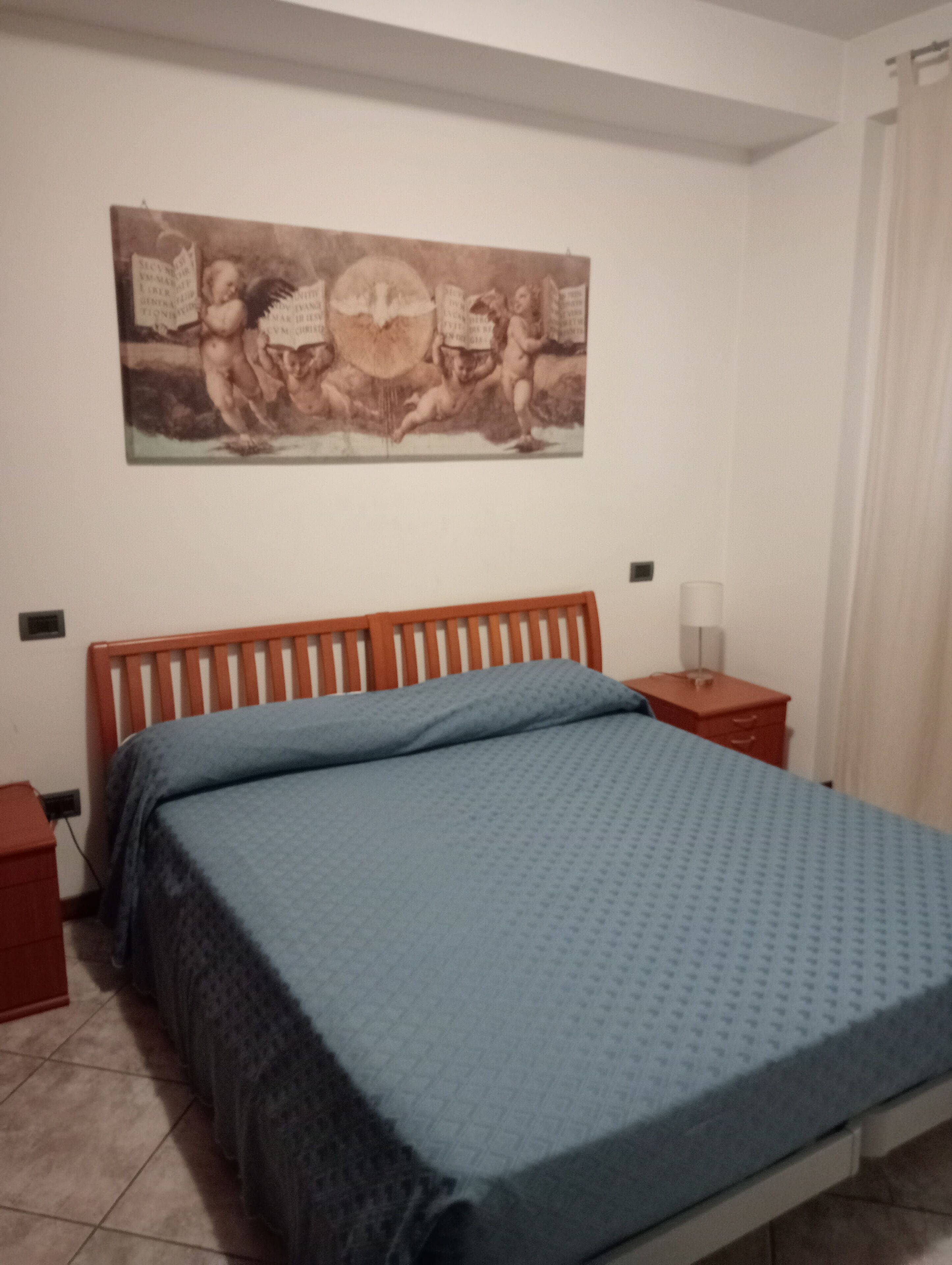 Ein Schlafzimmer mit einem Holzbett, einer blauen Bettdecke, einem Nachttisch mit Lampe und einem an der Wand hängenden Gemälde.