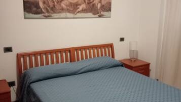 Ein Schlafzimmer mit einem Holzbett, einer blauen Bettdecke, einem Nachttisch mit Lampe und einem an der Wand hängenden Gemälde.