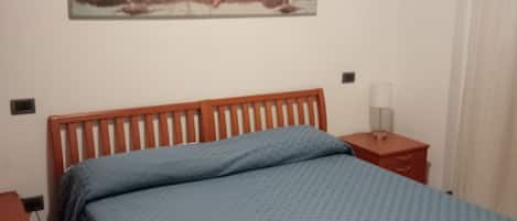 A bedroom with a wooden bed, a blue bedspread, a nightstand with a lamp, and a wall-mounted painting.