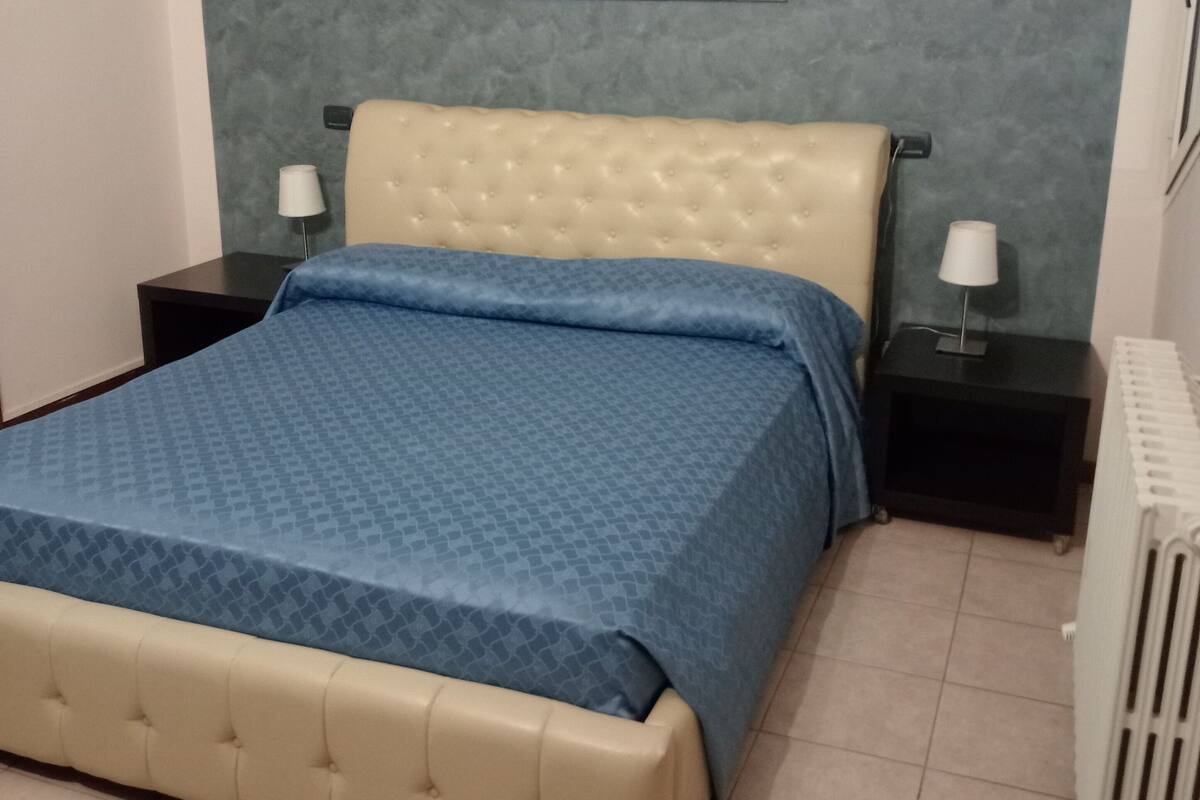 A bedroom with a bed, two bedside tables, a radiator, and a painting on the wall.