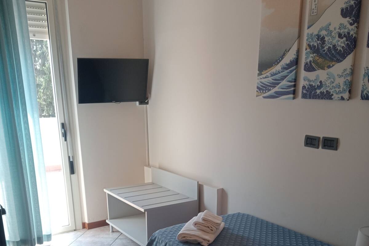 A bedroom with a bed, a desk, a TV mounted on the wall, and a window with curtains.
