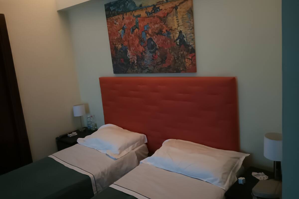 A hotel room with two beds, a red headboard, a painting on the wall, and a nightstand with a lamp.