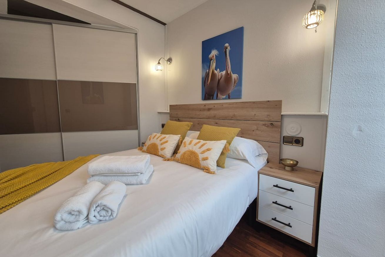 A neatly made bed with towels, a wooden headboard, a nightstand with drawers, a wall-mounted light fixture, and a painting of birds.