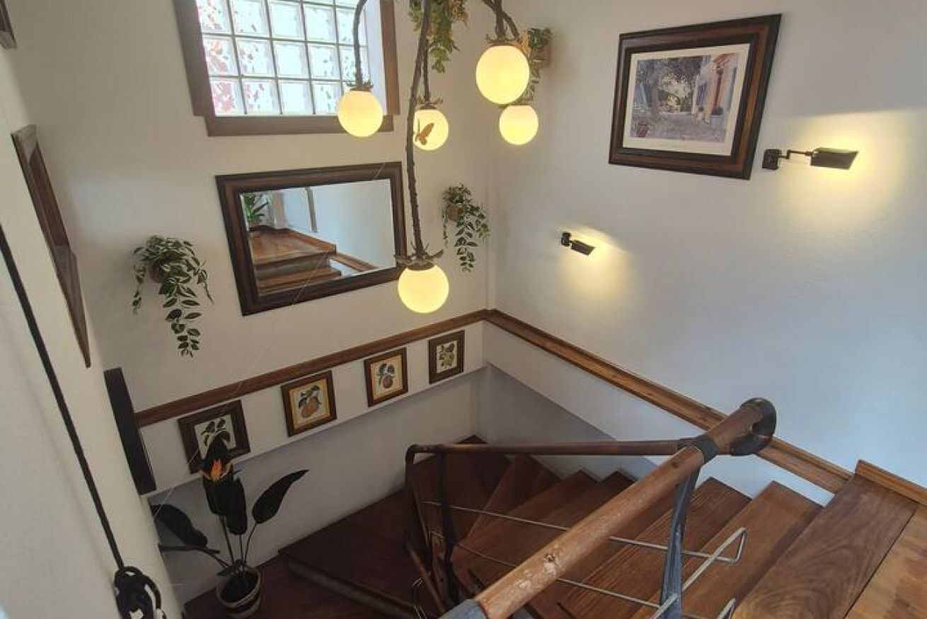 Una escalera con barandas de madera, una lámpara colgante, cuadros enmarcados y una ventana con vistas.