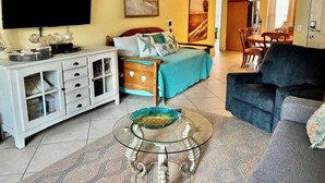 A living room with a sofa, a glass coffee table, a TV, a bed, and a dining area.