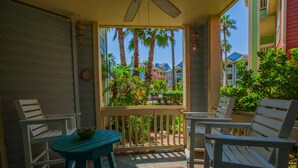 A balcony with two white chairs and a small table, overlooking a garden with palm trees and colorful buildings in the background.