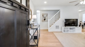 A modern living room with a dark wooden door, white walls, a staircase, a mounted TV, and a ceiling fan. - Charming 3BR Retreat in Chamblee (Chamblee)