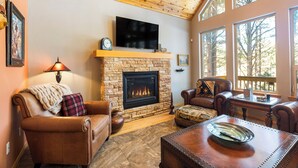 Una sala de estar acogedora con chimenea de piedra, muebles de cuero y ventanas amplias con vistas a árboles.