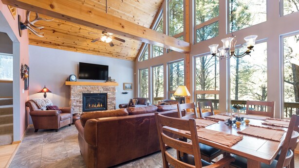 Un salón amplio con chimenea, sofás de cuero, una mesa de comedor con sillas y grandes ventanales que ofrecen vistas al bosque.