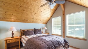 Un dormitorio con techo inclinado, una cama con cubrecama marrón, dos lámparas de noche y una mesita de noche con lámpara.