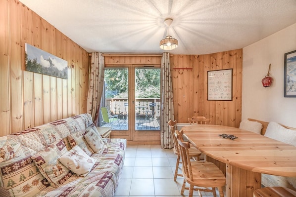 Una habitación acogedora con un sofá de madera, una mesa de comedor con sillas, una lámpara de techo y vistas a través de un ventanal.