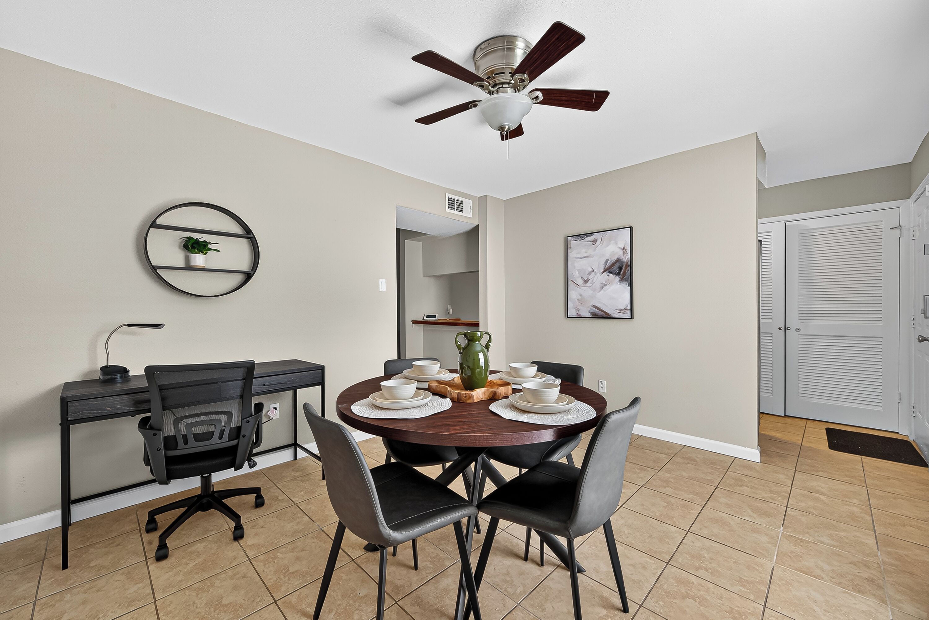 A dining area with a round table, four chairs, a ceiling fan, a wall-mounted shelf with a plant, and a door with a white closet.