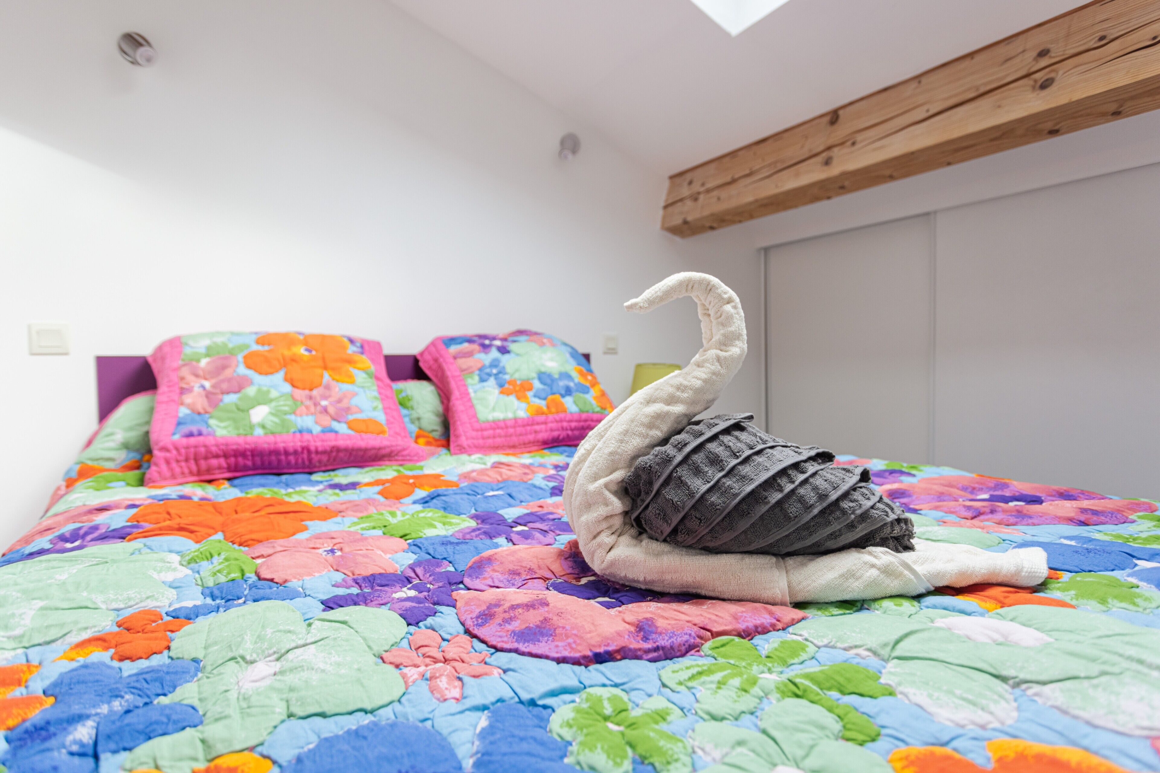 A bed with a colorful floral quilt, a swan-shaped pillow, and a wooden ceiling beam.