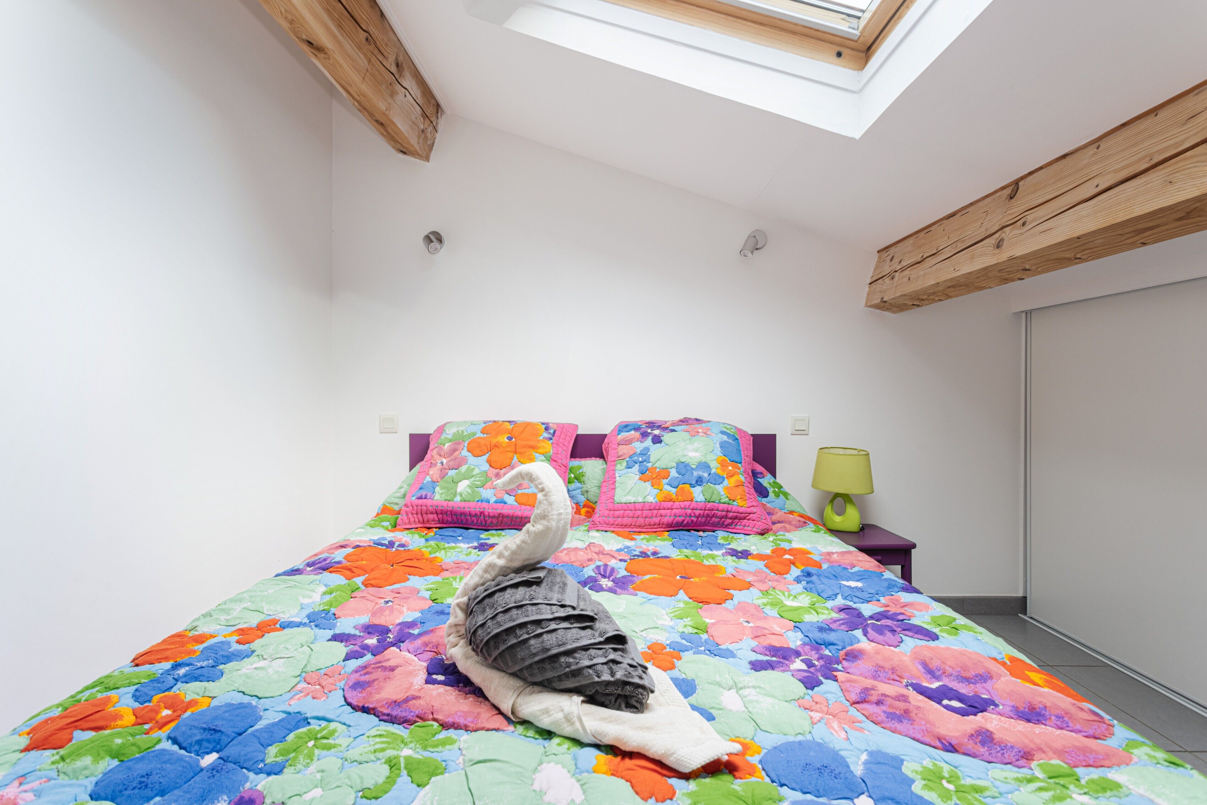 A bedroom with a skylight, a floral bedspread, and a bedside table with a lamp.