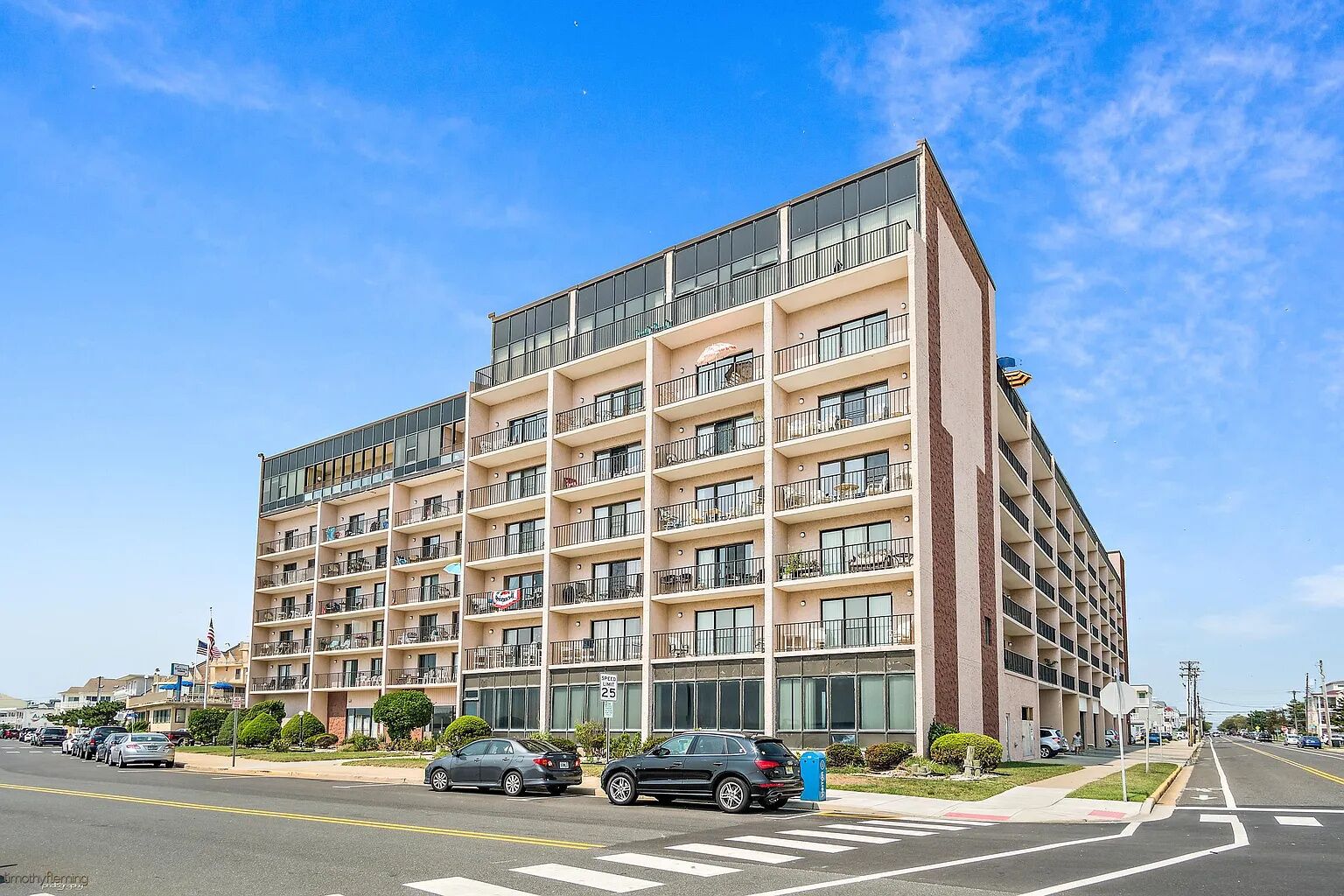 A multi-story hotel with a modern design, featuring balconies on each floor, a parking lot in front, and a clear blue sky.
