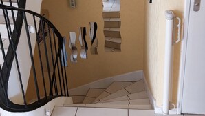 Un escalier en colimaçon avec une rampe noire, un luminaire fixé au mur et une porte dotée d’une poignée.