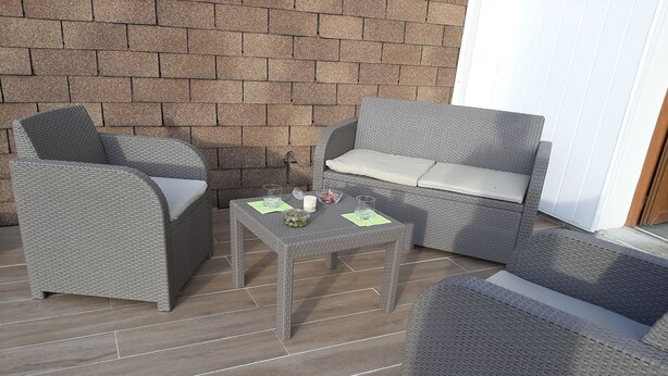 Un espace patio aménagé avec des meubles en osier, notamment un canapé et des fauteuils, ainsi qu’une petite table sur laquelle se trouvent un verre et un bol.