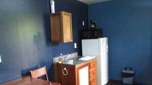 Een compacte keuken met een houten tafel en stoelen, een klein aanrecht met een spoelbak, en een witte koelkast.