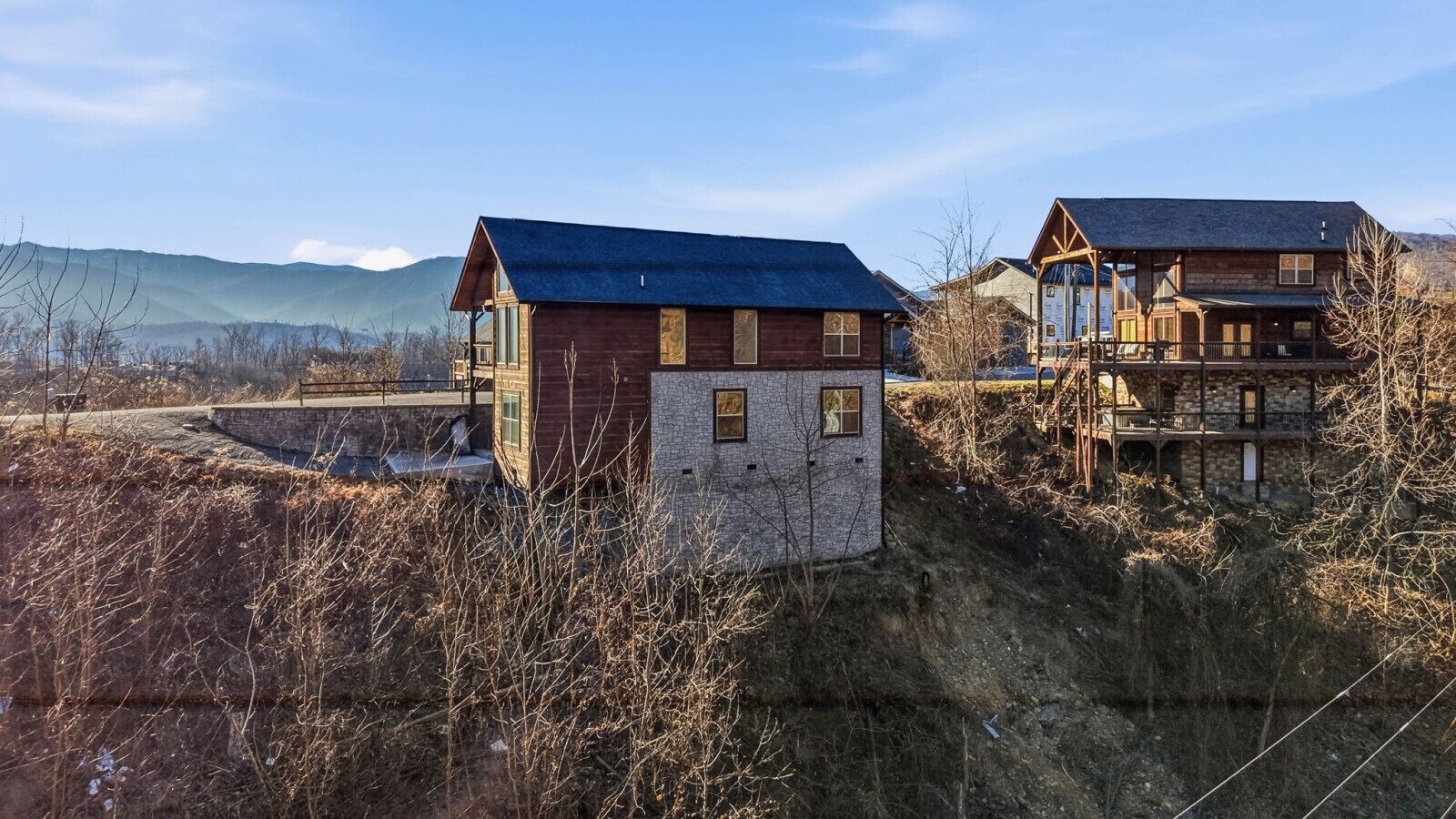 A rustic cabin with a stone foundation and a wooden upper structure, surrounded by dry vegetation and a mountainous backdrop.