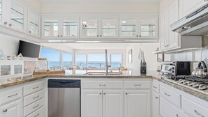 Una cocina moderna con armarios blancos, lavavajillas, una isla grande con pileta y vista al océano.