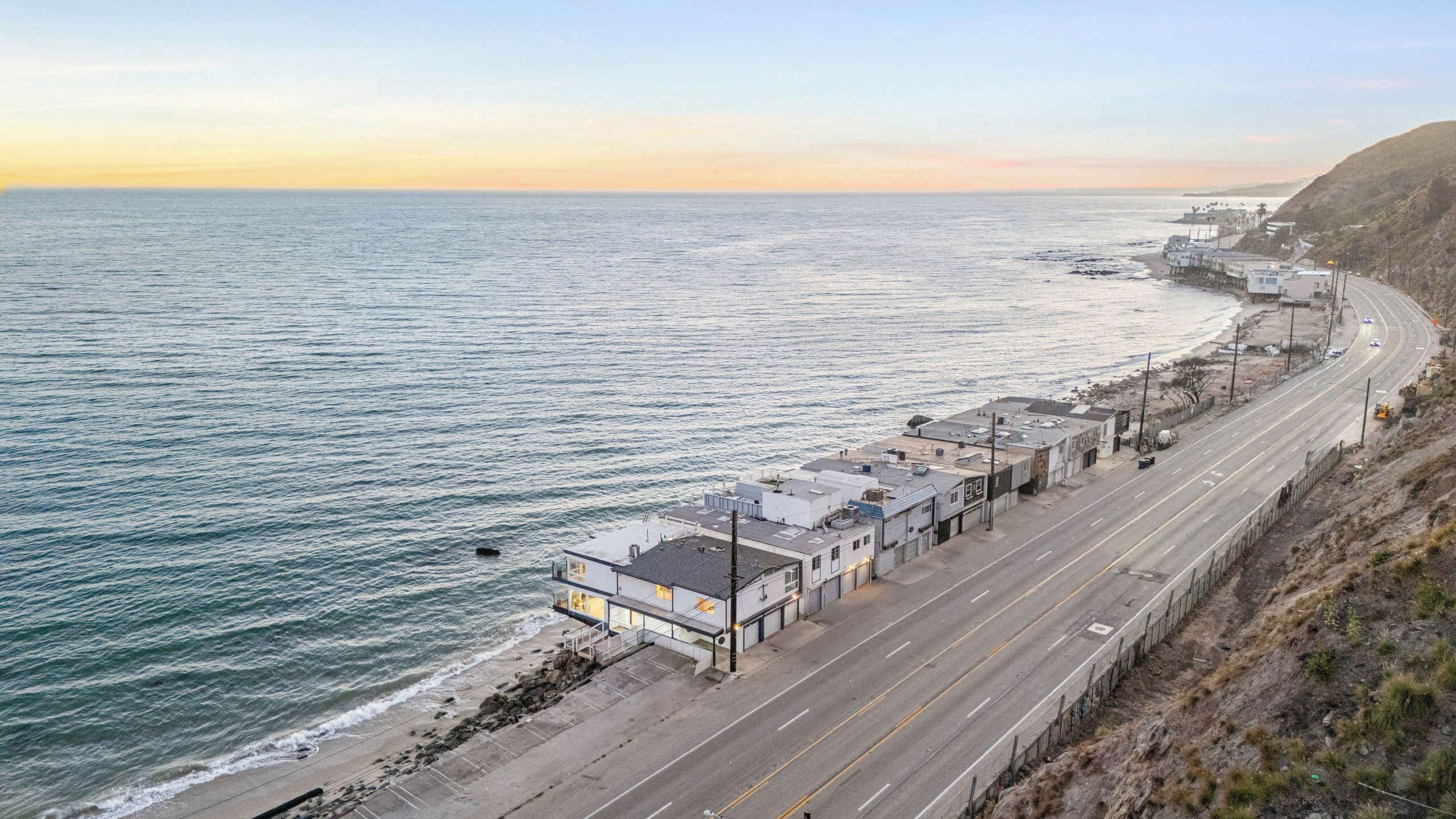 A coastal road with buildings, a beach, and a hilly landscape.
