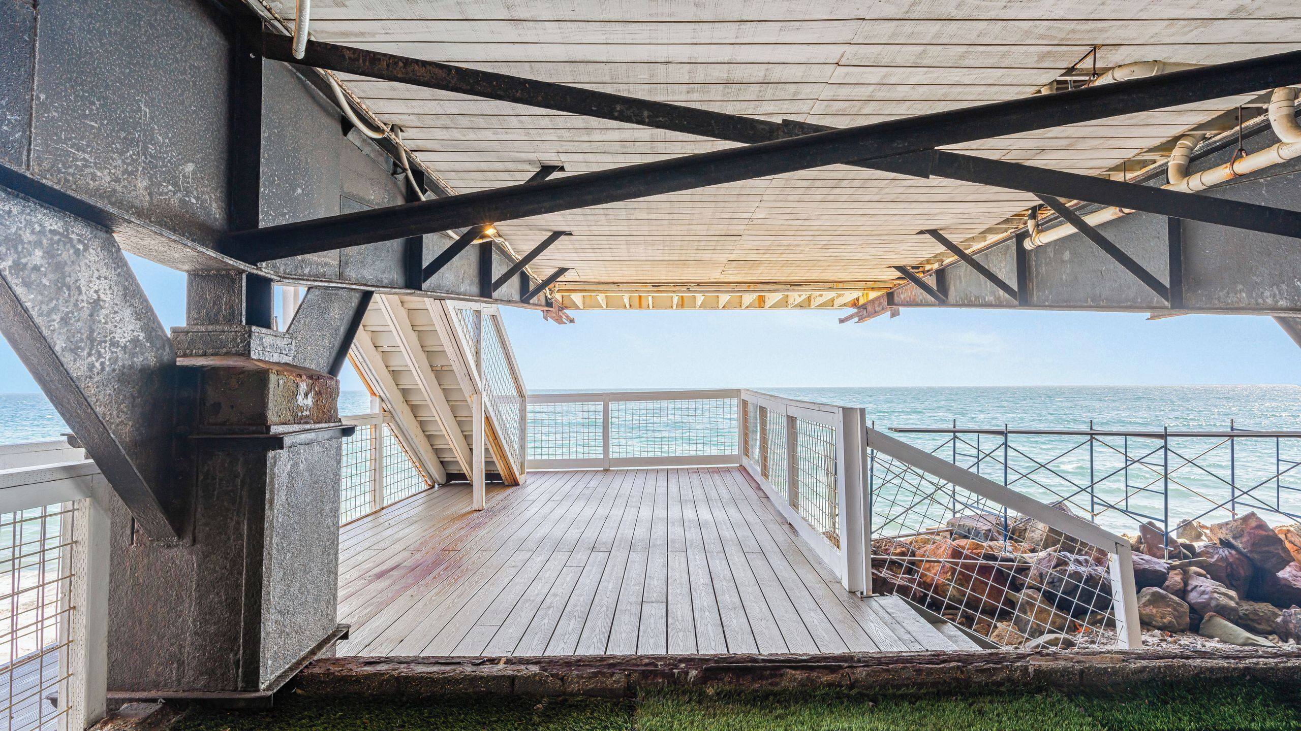 A wooden deck with a railing overlooking the sea, metal beams, and a view of the ocean.