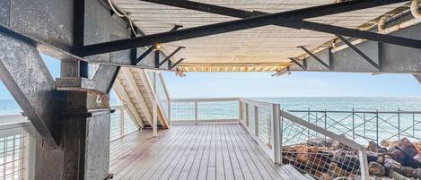A wooden deck with a railing overlooking the sea, metal beams, and a view of the ocean.
