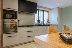A modern kitchen with built-in appliances, a dining table, and a window with curtains. - Chez neous – Welcoming Apartment in a Peaceful Farm Setting (Dommartin-lès-Remiremont)