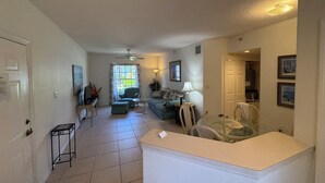 A spacious living area with a sofa, a small table, and a chair. There is a window with curtains, a ceiling fan, and a door leading to another room.